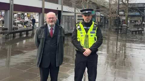 Swindon Borough Council Two men face the camera, one in a suit and tie, the other in a police uniform in a town centre with shops, benches, trees and a public bin in the background, which appears wet