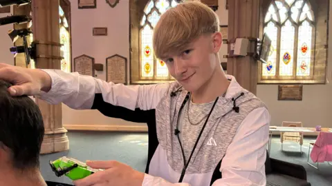 A 16 year old white boy with blonde hair is smiling at the camera while about to cut someone's hair to the left with barber razors. He is in a church in front of three stained glass windows. 
