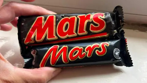 A Mars Bar from 1991 and a modern-day one held next to each other. The old Mars Bar is considerably bigger.