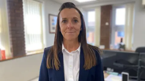 A woman with long brown hair smiles at the camera. She is in a school setting and is wearing a blue jacket and white shirt.