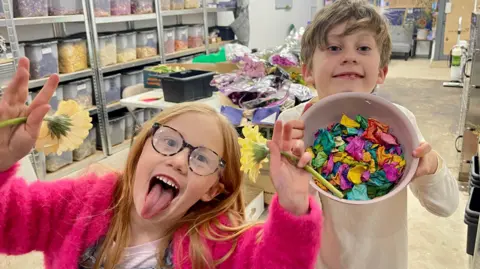 BBC A girl and boy stand in a shop unit. The girl on the left has long red hair, glasses, a pink fluffy cardigan, a pink t-shirt and denim dungarees. She is holding a flower in each of her raised hands and is sticking her tongue out. The boy has short fair hair, a long sleeved white top and is holding a bowl of bright confetti. Behind them are trestle tables covered in bunches and boxes of flowers. There are rows of shelves with labelled transparent boxes full of different coloured flower petals. The floor is bare concrete and the shop-front windows are covered with cardboard. 