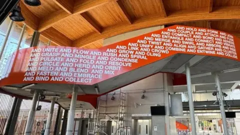 The interior of a new railway station, which still has some scaffolding up as it is not yet complete. The ceiling is wooden and beneath is a stairway balustrade, sweeping from left to right, which is coloured bright orange and has words written in white on it. 