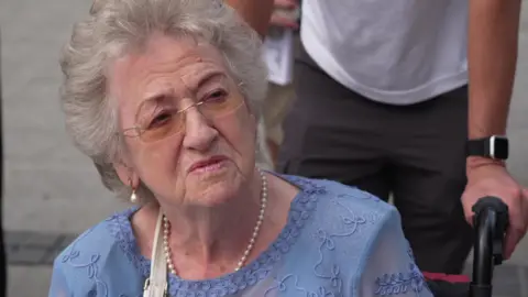 Frances is in a wheel chair and wears a blue top with a pearl necklace.