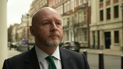 Liam Byrne wearing a suit with a green tie