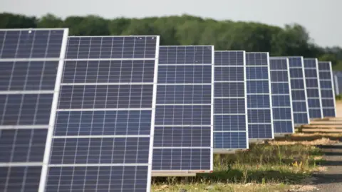 PA Media A general view of a solar farm. Rows of solar panels are placed in a field at a 45 degree angle.