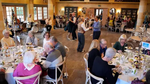 Groups of older people sit around round dining tables as they enjoy afternoon tea in a large dining room as people swing dance in the centre of the room