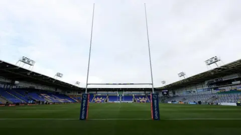 The inside of the Halliwell Jones Stadium, with pitch and goalposts and tiered seating in view.