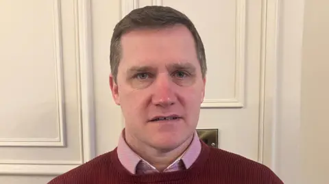 John McEntee pictured in a cream coloured room. He is wearing a red jumper with a pink shirt underneath. He has short light brown hair.