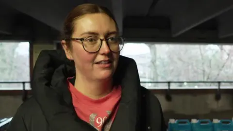 Sophie has tied back brown hair and dark framed glasses and is wearing a pink jumper and a black coat.