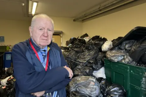 John Harrison standing next to a massive pile of bins. He has his arms crossed. He has thin, greying hair. He is wearing a blue shirt, tie and a blue jacket