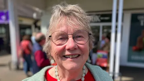 An older woman with glasses wearing a red top under light green coat smiles widely