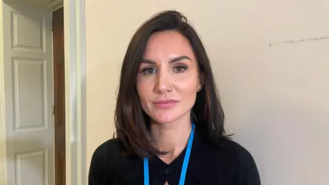Lisa McCartney is standing looking into the camera in front of a white wall. She is wearing a black top and a blue lanyard