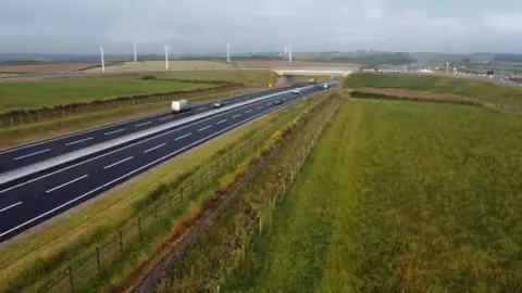 An aerial photo showing a stretch of the A30. There are wind turbines in the distance and a bridge across the dual carriageway. There are fields both sides of the road.