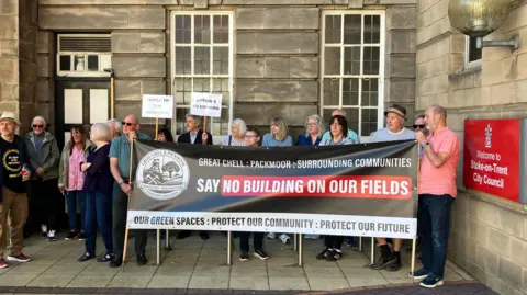 A group of campaigners, some holding signs on sticks, stand behind a rolled out banner which says "Say no building on our fields".
