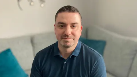 A man is sat on a sofa looking directly at the camera. He has dark hair and is wearing a blue top