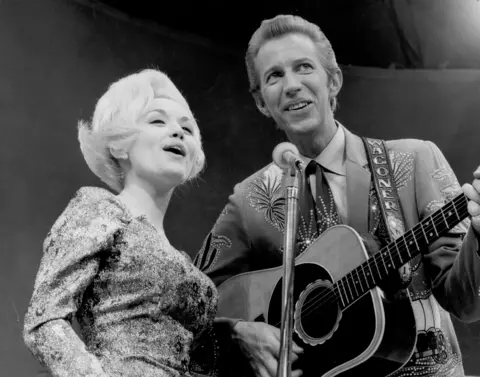 Michael Ochs Archives/Getty Images Dolly Parton and her collaborator Porter Wagoner perform onstage in circa 1967. She has a blond bob and a patterned dress and sings into the microphone with Porter, who plays guitar and is in a flashy suit and tie. 