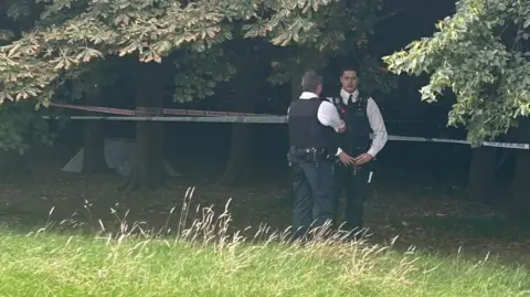 BBC Image of two officers talking in front of a cordon around tree trunks. A small white tent can be seen in the distance