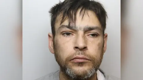A man with a greying beard and dark hair. he has brown eyes which are slightly narrowed
