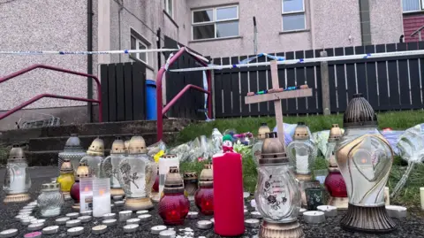 A collection of glass flashlights, candles, straps and a small wooden cross that were left near the block of floors where a man died after an assault in Dungannon. The floors are at the bottom and a white police cordon surrounds the building