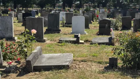 Phillip Norton/BBC Rows of gravestones in a cemetery. Several of them have been laid flat on the ground but the majority are upright. Each grave is separated by a grassed area. Shrubs and flowers are also visible.