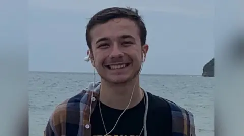 Family handout Aidan Webb with short dark hair smiling at the camera with the sea behind him.  He has earplugs around his ears and is wearing a multicoloured striped shirt.