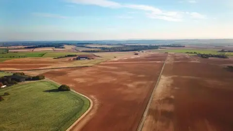 Andrew Wright An aerial view of a brown farm field with a concrete line down the middle indicating the location of the former Tarrant Rushton airfield