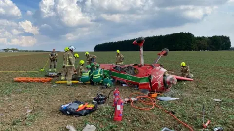 Essex Fire and Rescue Service  A red light aircraft upside down. Wheels are sticking up and it has been surrounded by emergency services including paramedics and firefighters