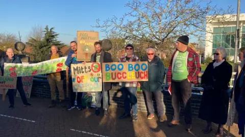 Laura Coffey/BBC Group of residents outside the council camber with (mainly) cardboard "save the wood" placards