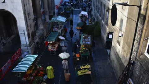 Bristol Film Image of Frog Lane. Cast members can be seen walking down the street, holding umbrellas. The set is a London market in the 1970s.