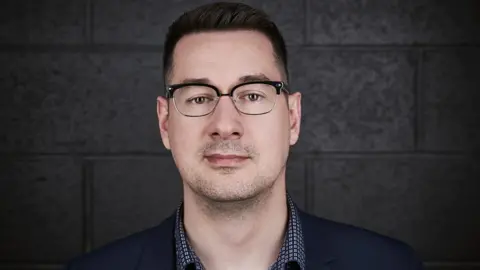 Behaviour Interactive Matthieu Côté stands in front of a dark grey brick wall. He looks impassive but happy. He's wearing a suit jacket with a gingham shirt underneath. He's got neatly cropped black hair and wears spectacles with thick black rims on the top half and wire rims at the bottom.