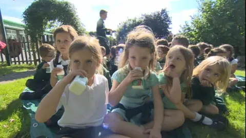 School children sitting outside on the grass on a sunny day drinking milk