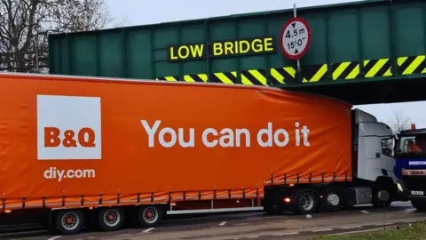 B&Q lorry bearing a logo that says 'you can do it' stuck under a bridge