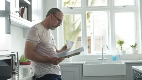 Getty Images Man standing in kitchen looking at a paper bill