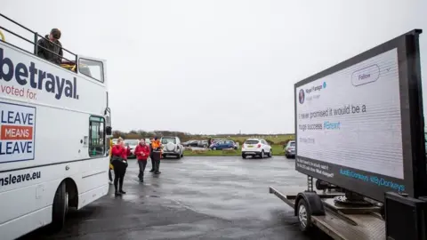Led By Donkeys Nigel Farage looking down from the top of a bus at the first leg of the March to Leave demonstration, with Led By Donkeys advertising van nearby.