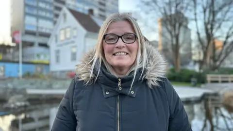 BBC A woman with blond hair and glasses smiles at the camera