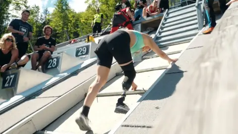 Red Bull  Milly Pickles near the finish line of the Red Bull 400 race. Milly is crawling up the steep incline, which is paved. She wears black shorts and a green-blue sports top. Her prosthetic leg is visible on her right and behind her spectators cheer her on.