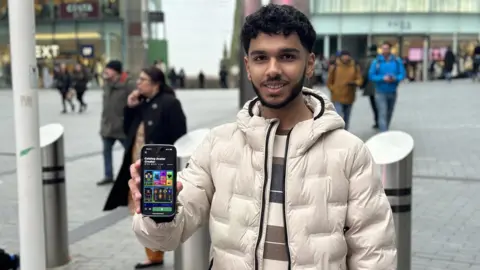 BBC A man wearing a beige puffa jacket stands in front of a shopping centre holding a phone showing a screen with the Roblox app on it