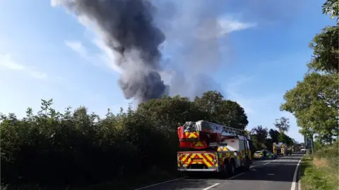 Smoke plume rising from behind a hedge. Fire engine in foreground