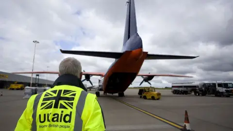 PA Media A UK aid staff member watches as cargo is loaded onto a plane