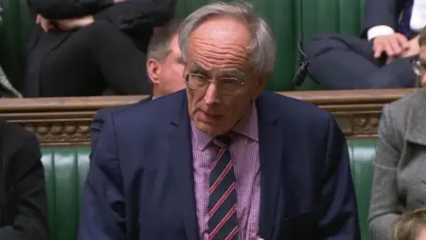 PA Media Man with grey hair and glasses, wearing a striped tie, speaks in the House of Commons