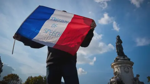 Getty Images A protester waves a French Tricolor flag with 'Freedom of Speech' written on it during an anti-terrorism vigil on Sunday