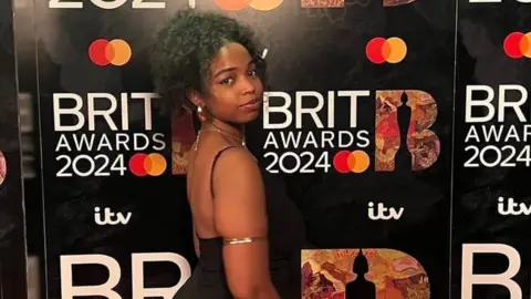 Nayana Brathwaite Nayana Brathwaite pictured on the Brits red carpet. Nayana is a 21-year-old black woman who wears her hair styled in an up do. She stands facing to the right, looking at the camera over her shoulder. She wears a strappy black dress and large gold earrings and arm bangle.