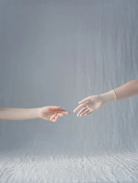 Kristina Varaksina Two hands reach for each other, one wearing a plastic glove