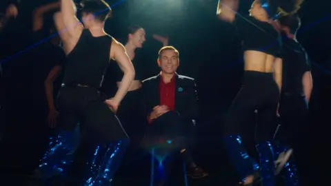 DHSC David Walliams sits among dancers in a still from the film to promte the Covid jab