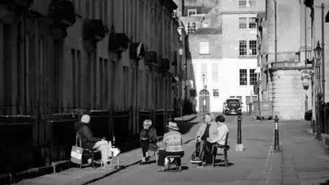 Jamie Bellinger Women sit apart from each other drinking champagne in street