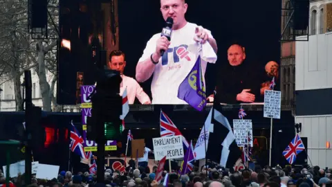 Getty Images Far-right activist Tommy Robinson seem on a screen addressing protesters gathered outside the Houses of Parliament