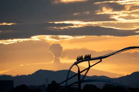 Guillermo Munoz / AFP A rollercoaster ride in Bogota with the Nevado del Ruiz volcano emitting a cloud of ash in the background