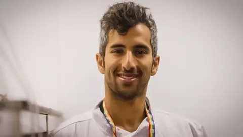 Submitted photos Numair Masud head and shoulder shot, smiling and wearing a rainbow flag lanyard