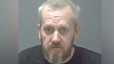 Suffolk Constabulary Mugshot of Peter Nash. It shows him with a brown and white beard. He has light grey hair and is wearing a dark navy t-shirt.