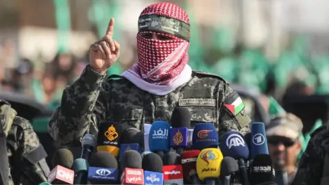Getty Images Abu Obeida speaks behind a number of microphones with slogans from the international media while wearing a scarf on his face, surrounded by Hamas fighters.